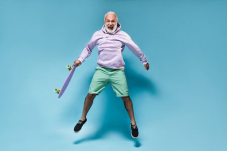 Веселый мужчина в толстовке прыгает со скейтбордом на синем фоне счастливый бородатый парень в фиолетовой одежде радостно подпрыгивает