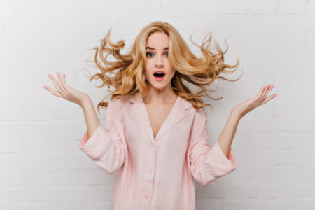 восторженная голубоглазая женщина с длинными светлыми волосами позирует перед белой кирпичной стеной, на которой запечатлена удивленная девушка в красивой розовой пижаме