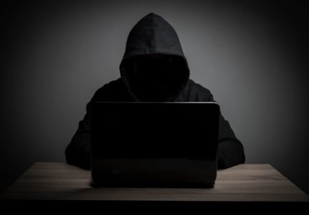человек в капюшоне с затемненным лицом сидит за столом работая на ноутбуке на темном фоне